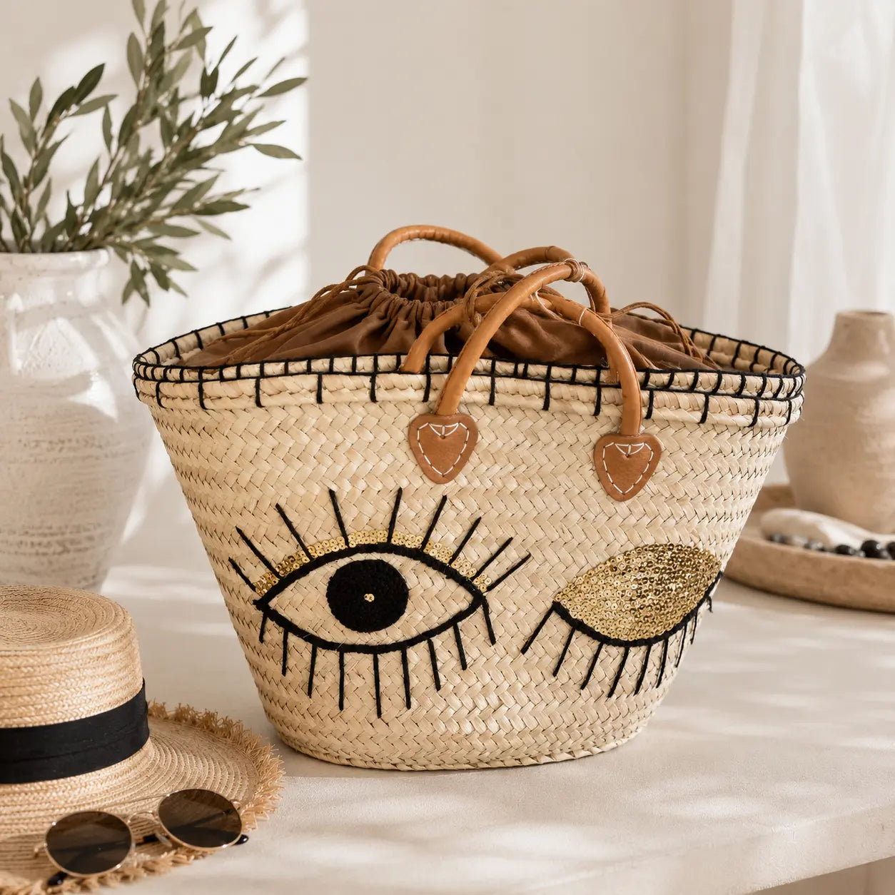 Grand panier bohème en paille naturelle avec motif yeux brodés noirs et sequins dorés, anses camel et fermeture intérieure à cordon, idéal pour la plage et l’été.