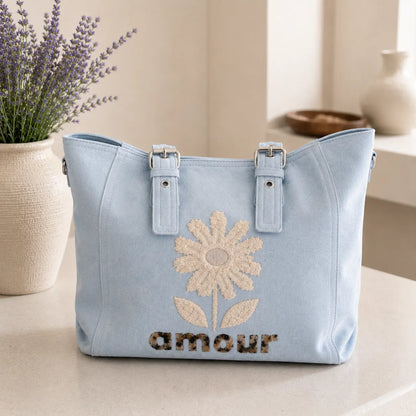 Sac cabas femme effet suédine avec fleur en relief et inscription Amour, disponible en bleu pastel et rose poudré, grande capacité avec multiples poches zippées.